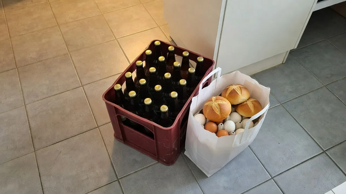 Eine rote Bierkiste mit 20 braunen Halbliterflaschen steht auf dem Küchenboden neben einer Papiertasche mit Eiern und Kaisersemmeln.